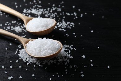 Photo of Organic salt in spoons on black wooden table, closeup. Space for text