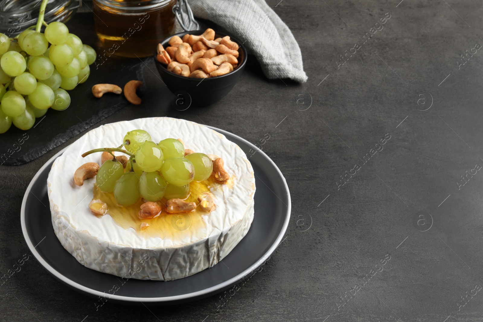Photo of Brie cheese served with grape, cashew nuts and honey on dark grey table. Space for text
