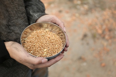 Poor homeless woman with bowl of wheat outdoors, closeup