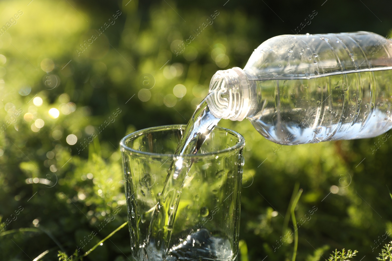 Photo of Pouring fresh water from bottle into glass on green grass outdoors, closeup