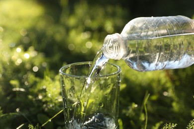 Pouring fresh water from bottle into glass on green grass outdoors, closeup