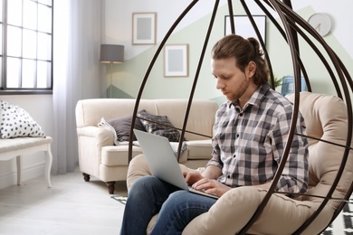 Photo of Young man working with laptop on hanging armchair in home office