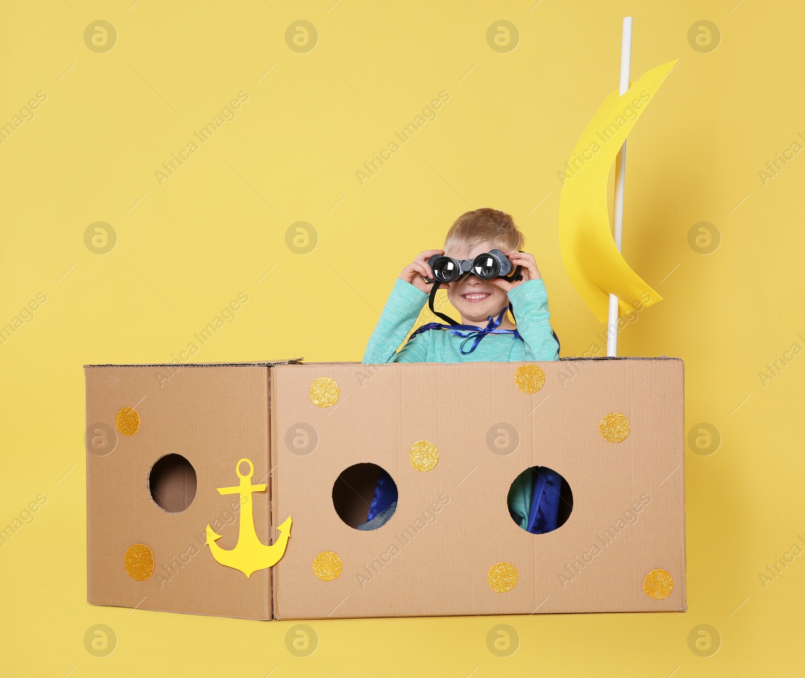 Photo of Little child playing with ship made of cardboard box on yellow background