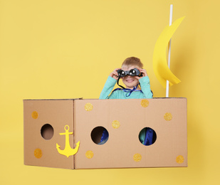 Photo of Little child playing with ship made of cardboard box on yellow background