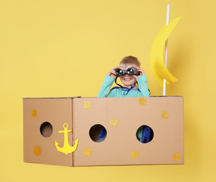 Little child playing with ship made of cardboard box on yellow background