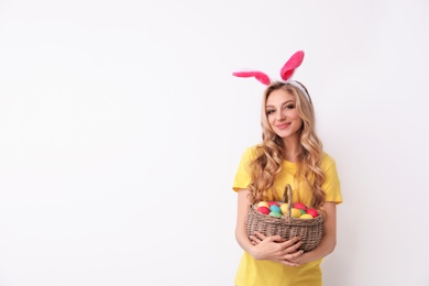 Photo of Beautiful young woman in bunny ears headband holding basket with Easter eggs on light background, space for text