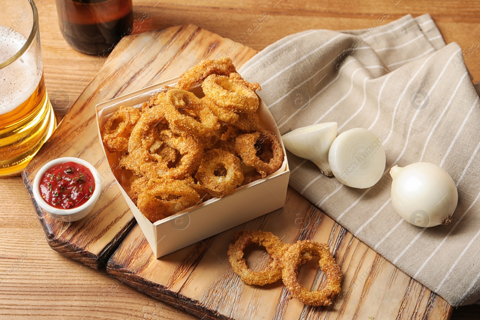 Photo of Cardboard box with crunchy fried onion rings and tomato sauce on wooden board