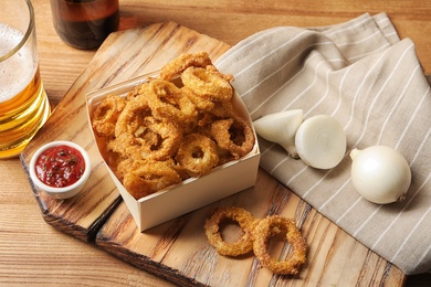 Photo of Cardboard box with crunchy fried onion rings and tomato sauce on wooden board