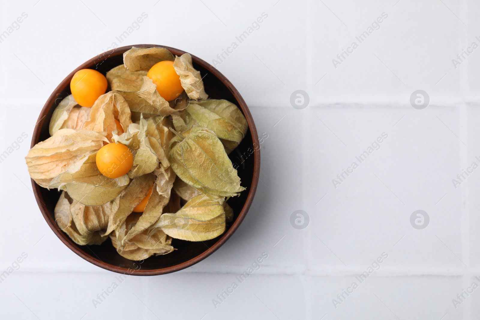 Photo of Ripe physalis fruits with calyxes in bowl on white tiled table, top view. Space for text