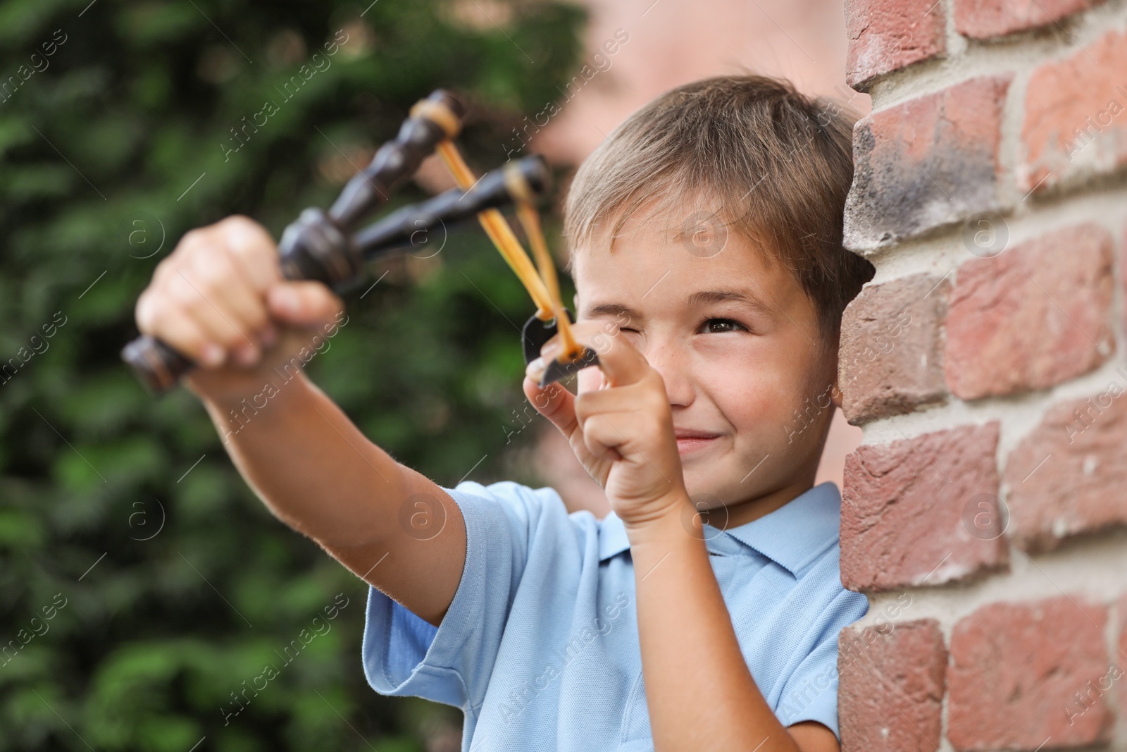 Photo of Little boy playing with slingshot near brick wall outdoors