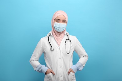Photo of Muslim woman wearing hijab, medical uniform and protective mask on light blue background