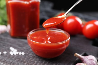 Taking tasty ketchup with spoon from bowl at black wooden table, closeup. Tomato sauce