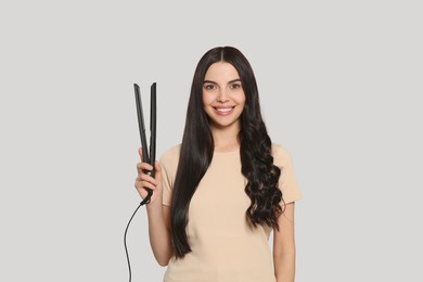 Beautiful happy woman with hair iron on light grey background