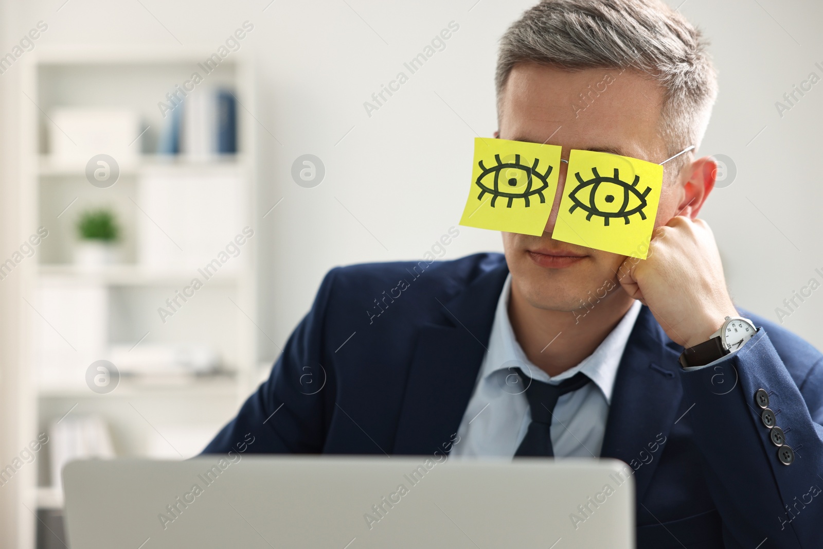 Photo of Man with fake eyes painted on sticky notes snoozing at workplace in office