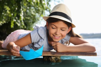 Cute little girl playing with paper boat on wooden pier near river