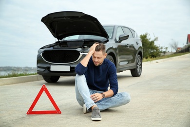 Upset man sitting near warning triangle and broken car on road
