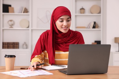 Photo of Muslim woman in hijab using laptop at wooden table in room