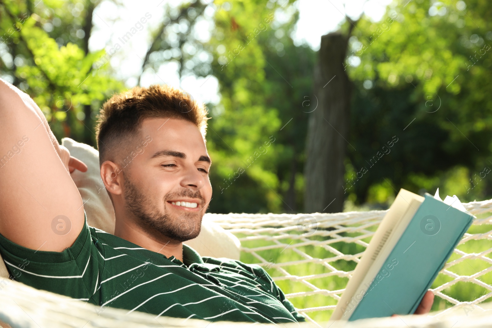 Photo of Young man reading book in comfortable hammock at green garden