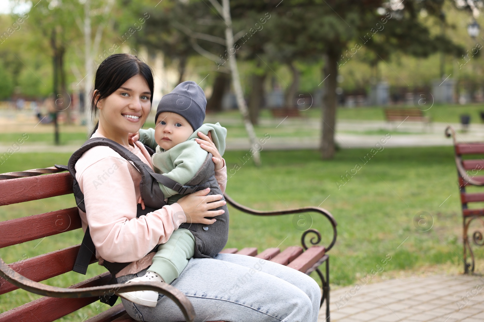 Photo of Mother holding her child in sling (baby carrier) on bench in park. Space for text