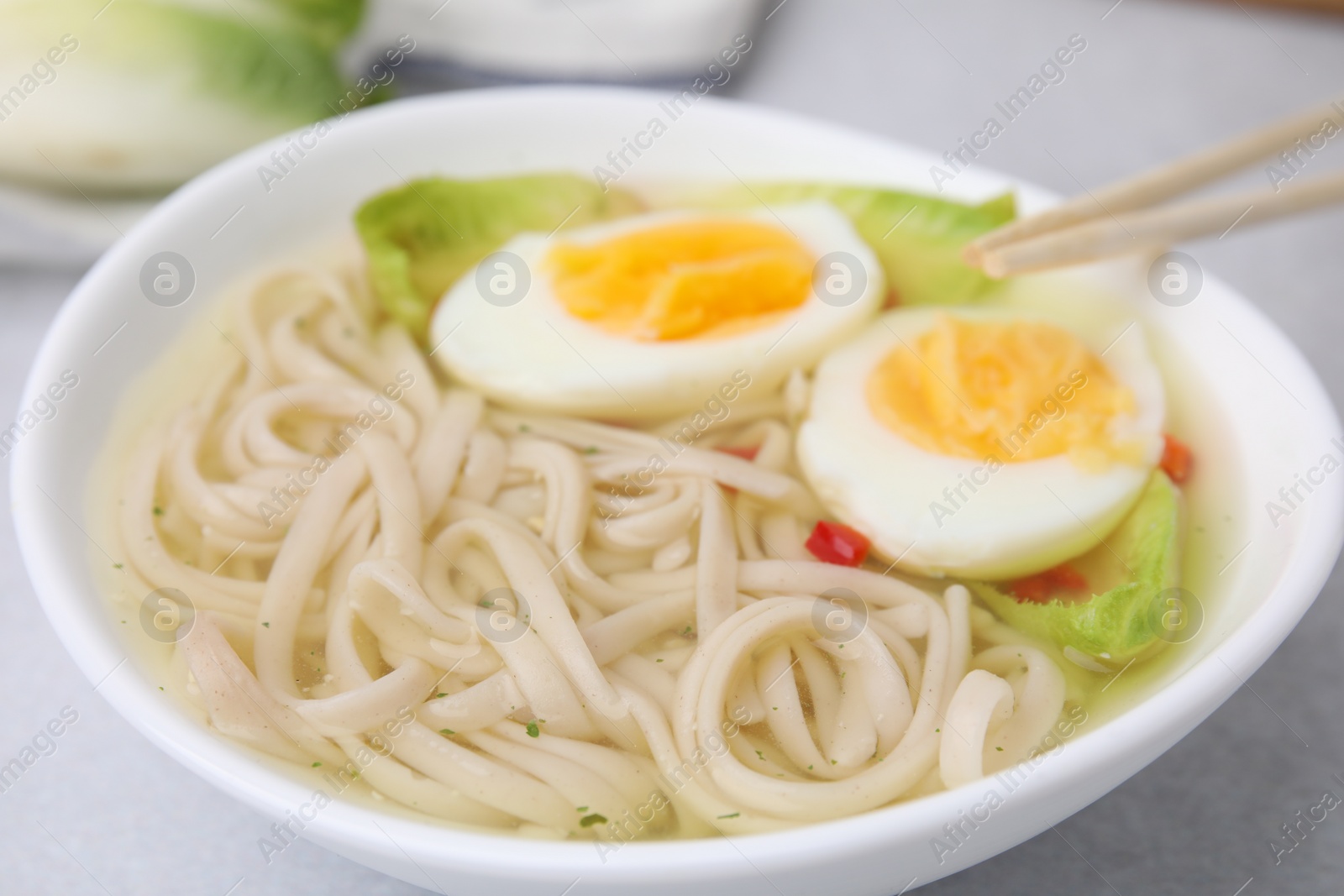 Photo of Bowl of delicious rice noodle soup with celery and egg on light grey table, closeup