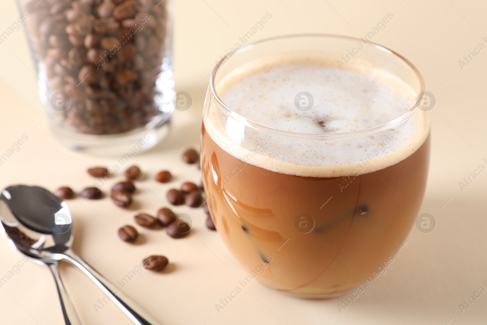 Photo of Refreshing iced coffee with milk in glass, beans and spoons on beige table, closeup