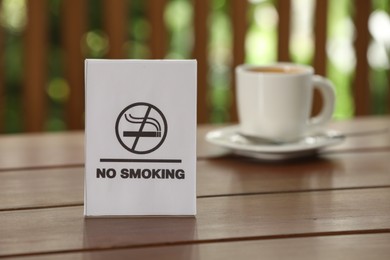 Photo of No Smoking sign and cup of drink on wooden table against blurred background, selective focus