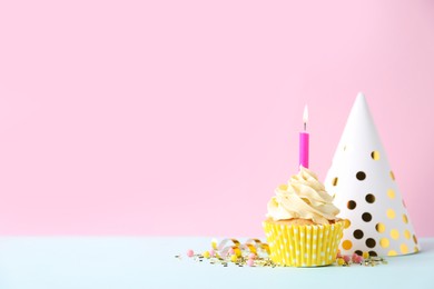 Delicious birthday cupcake with burning candle and party decor on white table against pink background, space for text