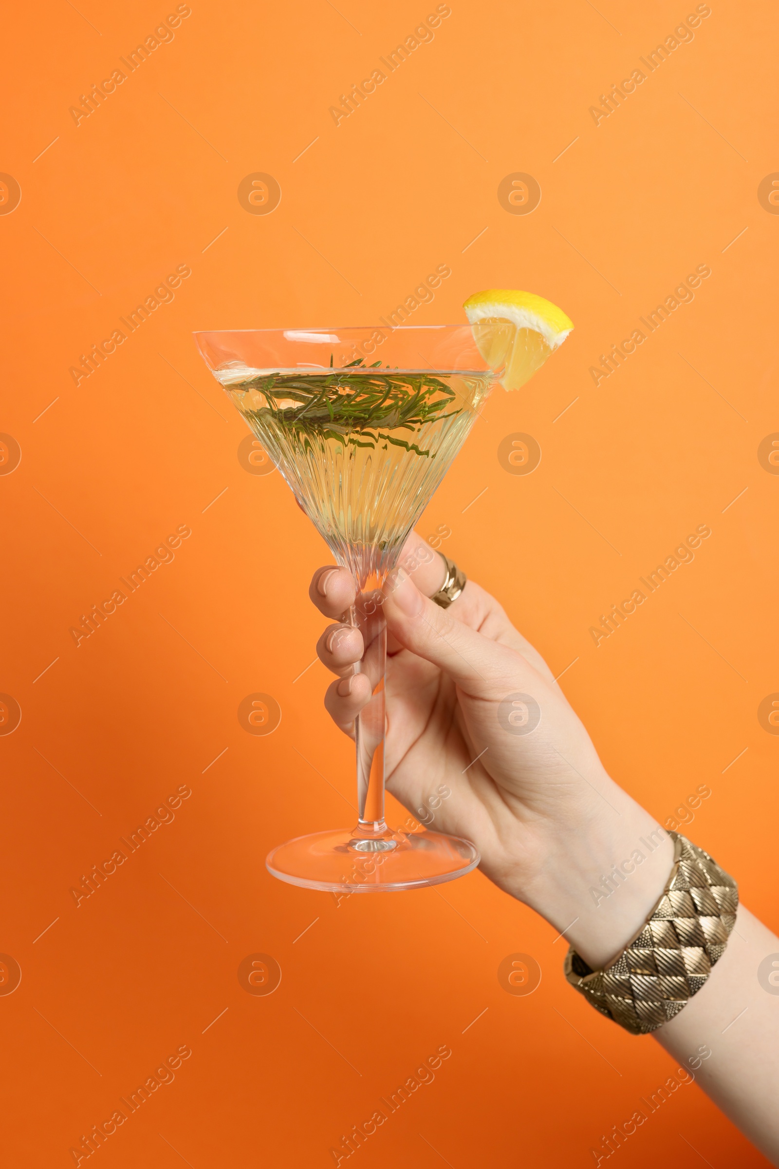 Photo of Woman holding martini glass of refreshing cocktail with lemon slice and rosemary on orange background, closeup