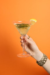 Woman holding martini glass of refreshing cocktail with lemon slice and rosemary on orange background, closeup