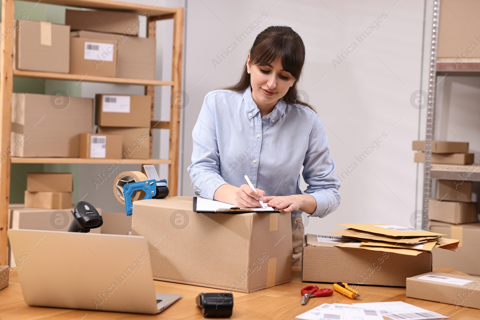 Photo of Parcel packing. Post office worker with clipboard writing notes at wooden table indoors