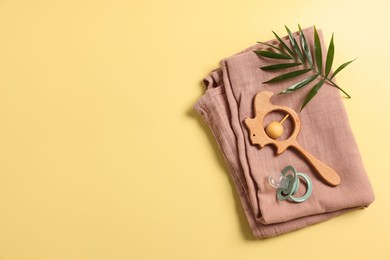 Baby accessories. Wooden rattle, pacifier, palm leaf and cloth on yellow background, top view. Space for text