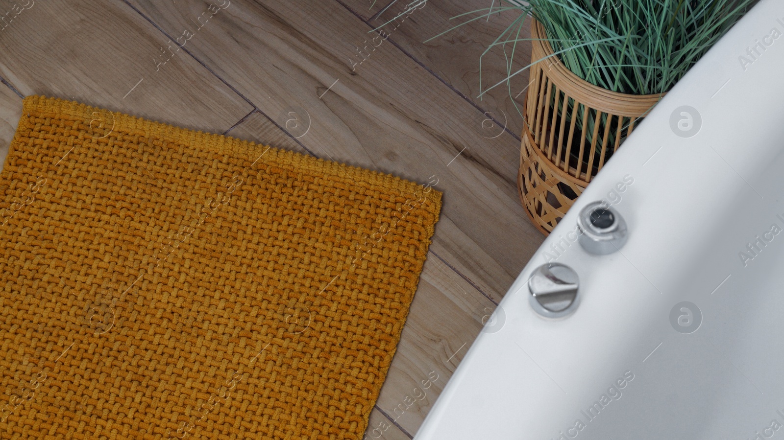 Photo of Soft mustard color bath mat near tub and houseplant on wooden floor in bathroom, above view