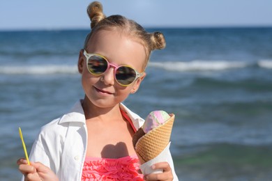 Adorable little girl with delicious ice cream near sea on sunny summer day, space for text