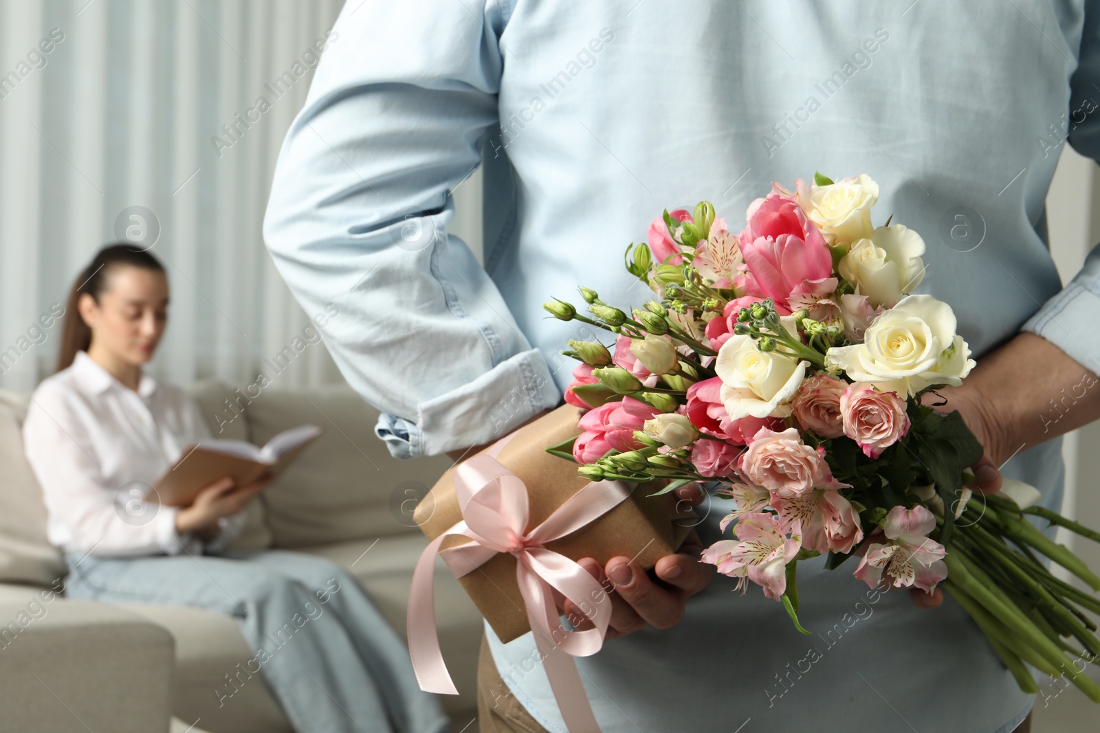 Photo of Man hiding bouquet of flowers and present for his beloved woman indoors, closeup