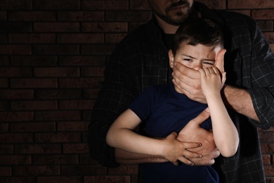 Photo of Adult man covering scared little boy's mouth near brick wall, space for text. Child in danger