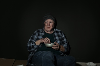 Poor senior man with bread and bowl on floor near dark wall