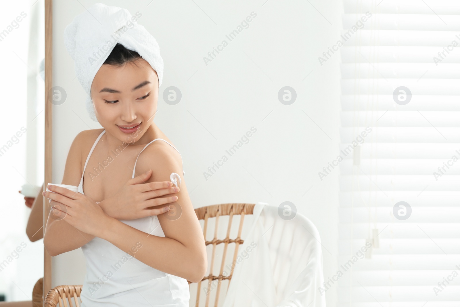 Photo of Beautiful young Asian woman applying body cream on shoulder in room, space for text