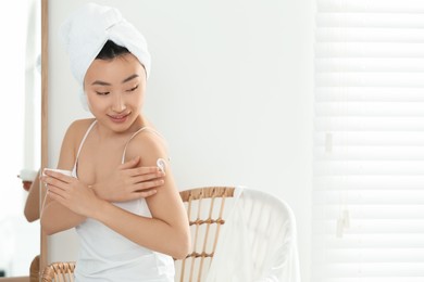 Beautiful young Asian woman applying body cream on shoulder in room, space for text