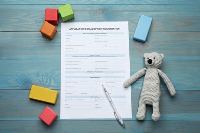 Photo of Adoption application, toy bear, colorful cubes and pen on blue wooden table, flat lay