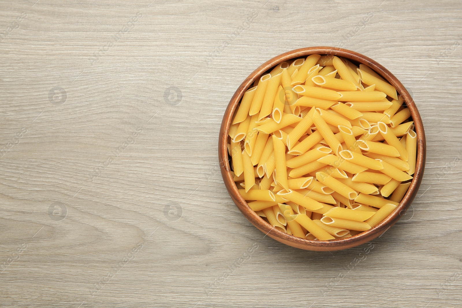 Photo of Raw penne pasta in bowl on light grey wooden table, top view. Space for text