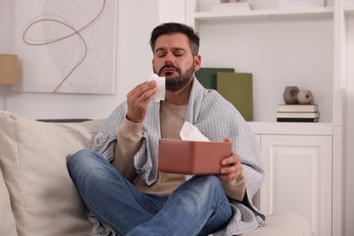 Photo of Sick man wrapped in blanket with tissues on sofa at home. Cold symptoms