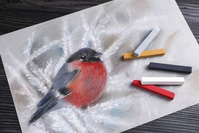 Photo of Colorful chalk pastels and beautiful painting of bullfinch on black wooden table, top view