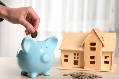 Photo of House model and money on wooden table. Woman putting coin into piggy bank indoors, closeup