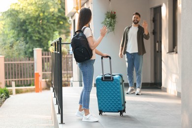 Photo of Long-distance relationship. Man waving to his girlfriend with luggage near house entrance outdoors, selective focus