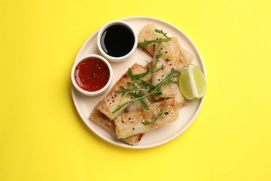 Photo of Tasty fried spring rolls with arugula, lime and sauces on yellow table, top view