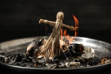 Photo of Voodoo doll burning in metal tray on dark background. Curse ceremony