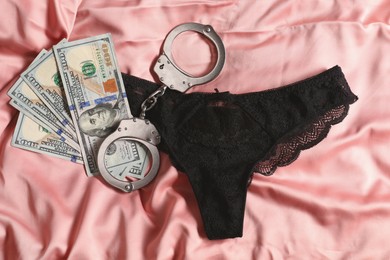 Prostitution concept. Black panties, dollar banknotes and handcuffs on pink silk fabric, flat lay