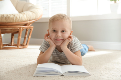 Photo of Cute little boy reading book on floor at home
