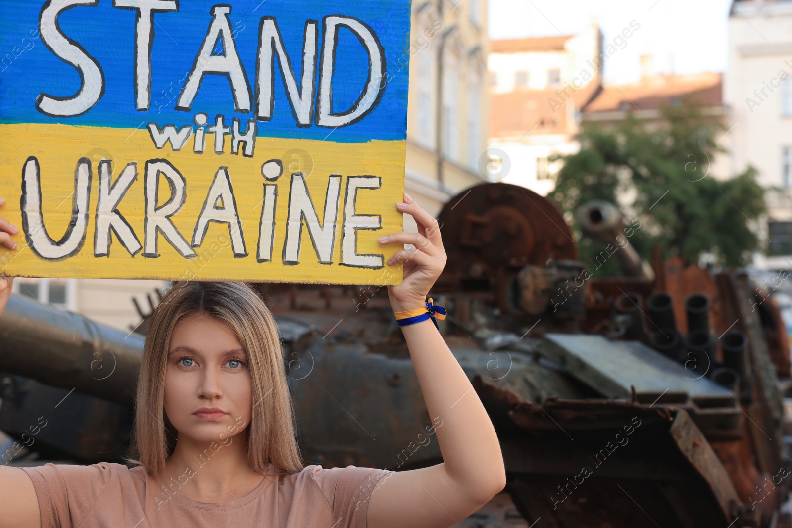 Photo of Sad woman holding poster in colors of national flag with words Stand with Ukraine near broken tank on city street