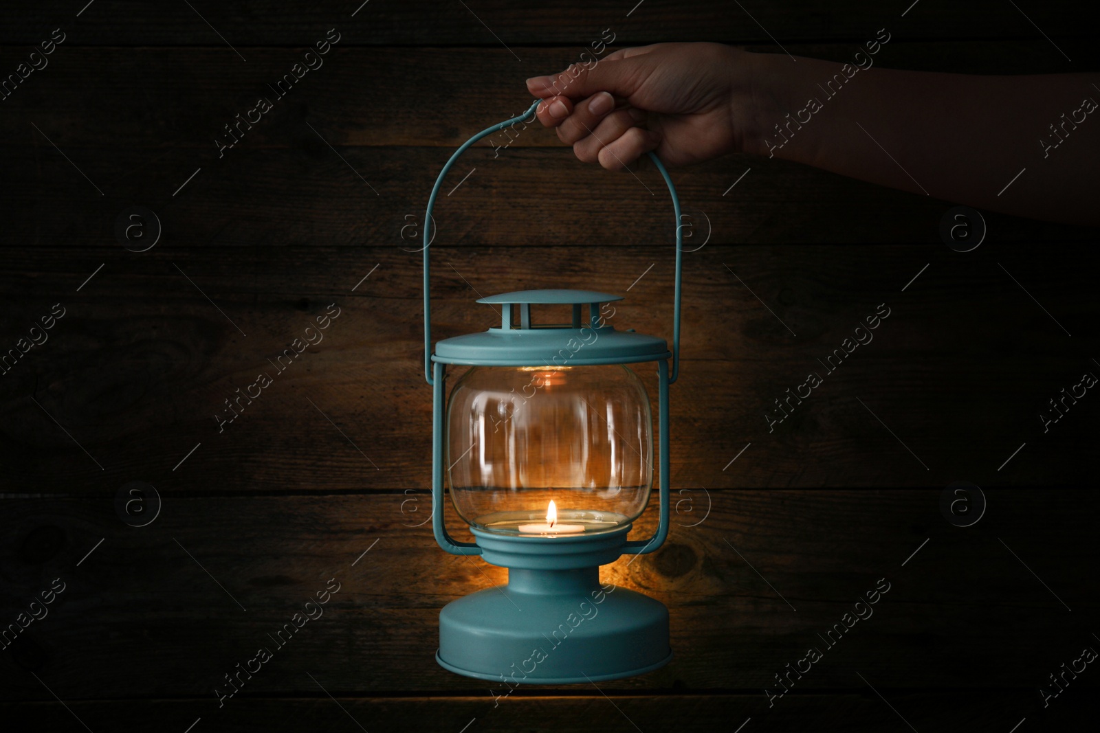 Photo of Woman holding lantern with burning candle against wooden wall in darkness, closeup